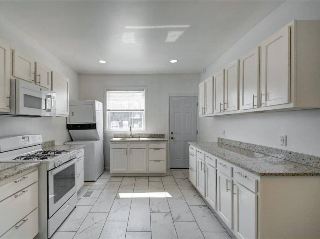 Renovated galley kitchen with white cabinets, granite counters, gas range, and stacked laundry